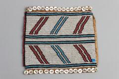 Beaded patch with white background with stripes. Four stripes of blue and black beads. Twelve diagonal shorter stripes of black, blue and red. Red bead trim on one end. Button trim on two ends. One round blue tack at one corner. Leather border on back, white tag attached.