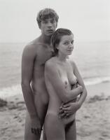 A photograph of a nude man and woman standing on the beach. The man stands behind the woman, wrapping his arm around her waist. They both look into the camera lens.