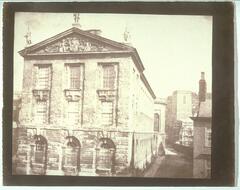 This photograph depicts a view of an old building with three floors of tall, grated windows, a frieze near its roof, topped with three statues. A street runs down the side of the building where there is a church with scaffolding on it.