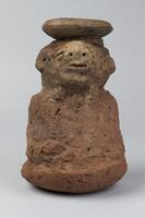 Red clay vessel in the form of a rounded cylinder with the upper portion in the form of a face. The eyes are cowrie shells set into the clay, while the nose is broad and the mouth underneath is thin. Small semi-circular ears protrude from the each side of the face. The vessel is topped by a circular rim, the back portion of which is missing. 