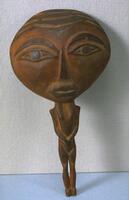 A wooden spoon that is shaped like a human. The handle is the body with two legs and two arms that are crossed in from of the body, hands clasped. The backside of the bowl of the spoon has a face.