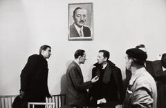 A photograph of men in suits meeting in a room with white walls on which a portrait of a man hangs.
