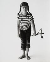 Girl sticking out her tongue, holding a slingshot, barefoot and waring a striped shirt.
