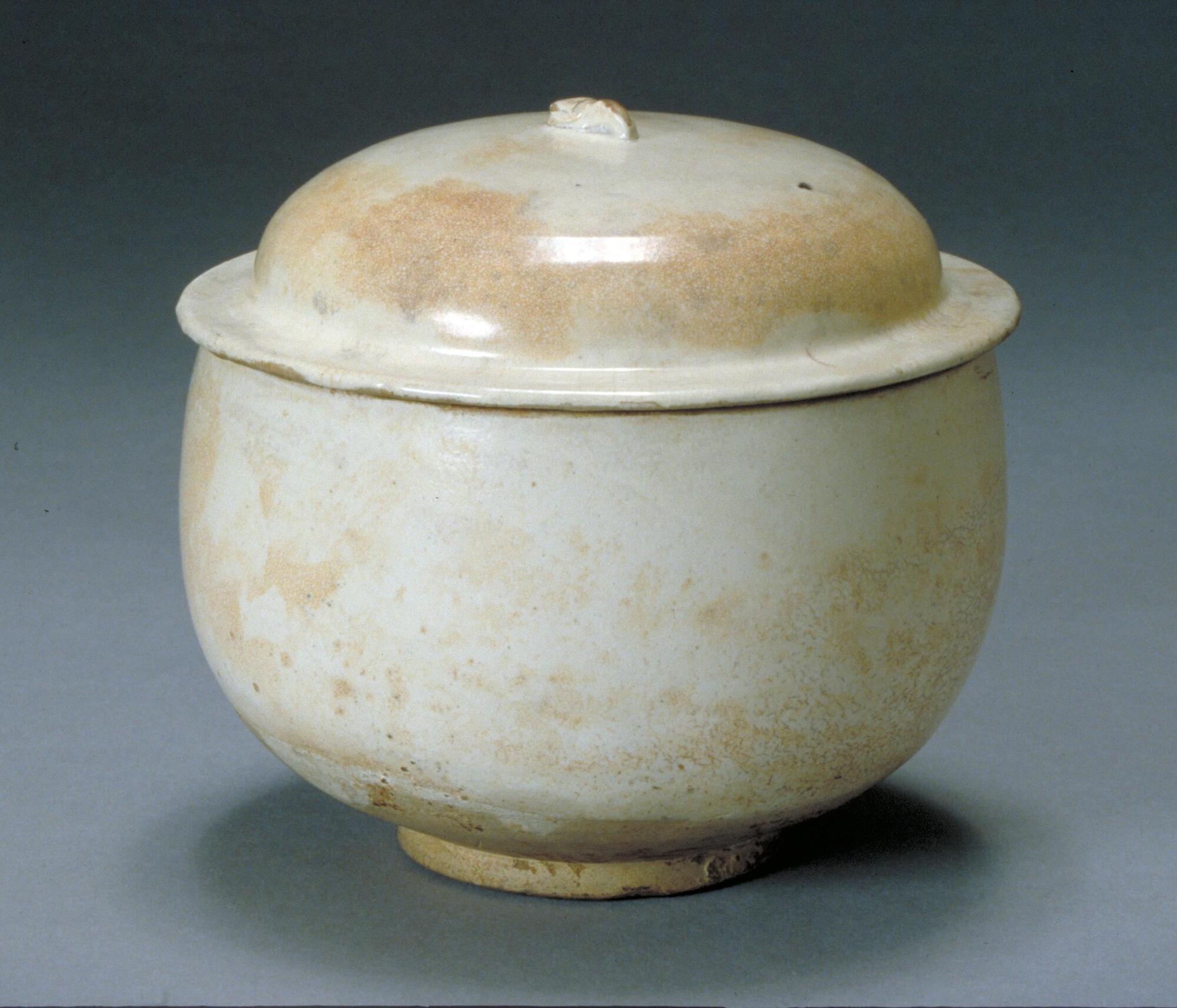A tall, round bowl with a foot at the bottom and a lid that starts out flat but then becomes a rounded top. It is white with some yellow coloring.