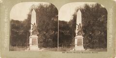 This black and white stereoscopic image features two images of that Boston Massacre Monument. A sculpture of a man holding a spear in front of a large, vertical, white, piller with a pointed top. Trees take up the right side of the image.