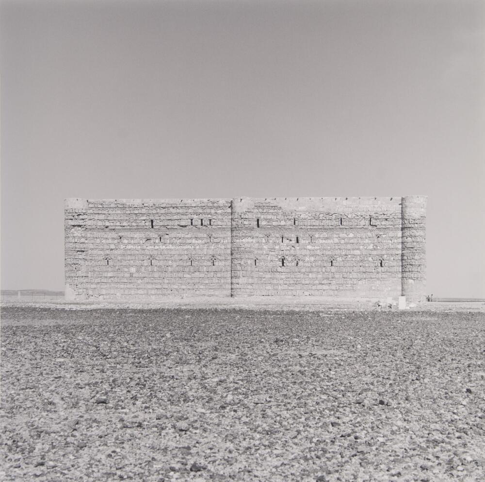 The outer walls of Karanah Castle in Jordan. The desert ground makes up the entire foreground with the horizon line almost reaching the half-point of the photograph. Just below the horizon is the base of the structure. The castle itself is a rectangular brick wall with three rounded pillars built into it, two at the corners and one in the center. The sky above it is clear and cloudless.