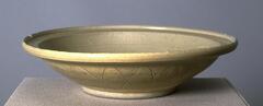 A wide, shallow stoneware bowl on a footring with an everted rim with articulation.  The exterior is carved with lotus petals and covered in a gray-green celadon glaze.