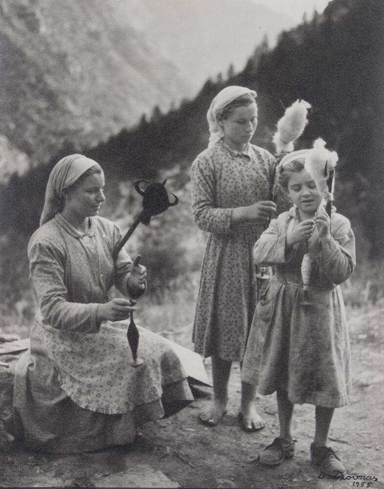 In this photograph, three young women of varying ages use drop spindles to spin wool in a mountainous setting. The eldest of the three is seated in the left foreground and appears to monitor her younger companion on the opposite side of the photograph. All three figures wear unadorned, patterned and/or plain dresses with their hair wrapped in cloth coverings.&nbsp;