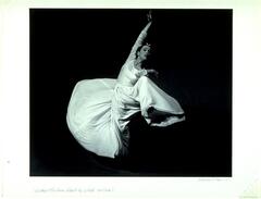 A photograph of a woman dancing. She wears a dress with flowing fabric, creating a sense of motion as she performs a jump.