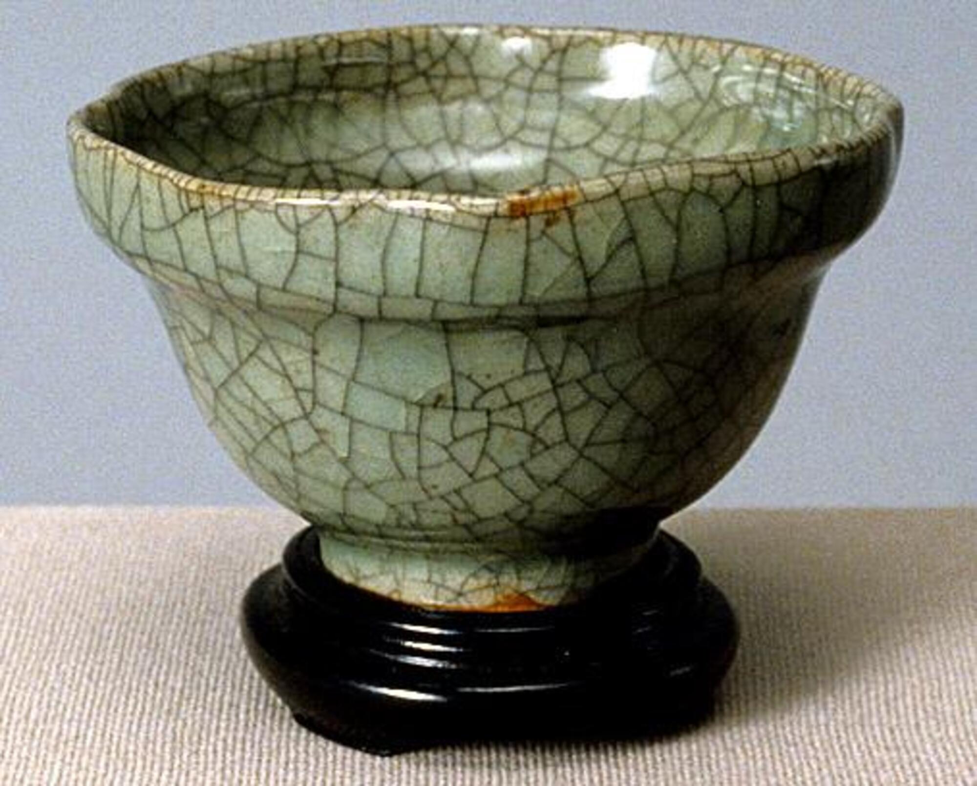 A stoneware, bowl-shaped cup on a tall foot ring with an articulated five-lobed rim. It is covered in a crackled celadon glaze. 