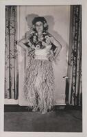 A woman standing with her hands on her hips, dressed in a grass skirt and wearing flowers around her neck and in her hair.