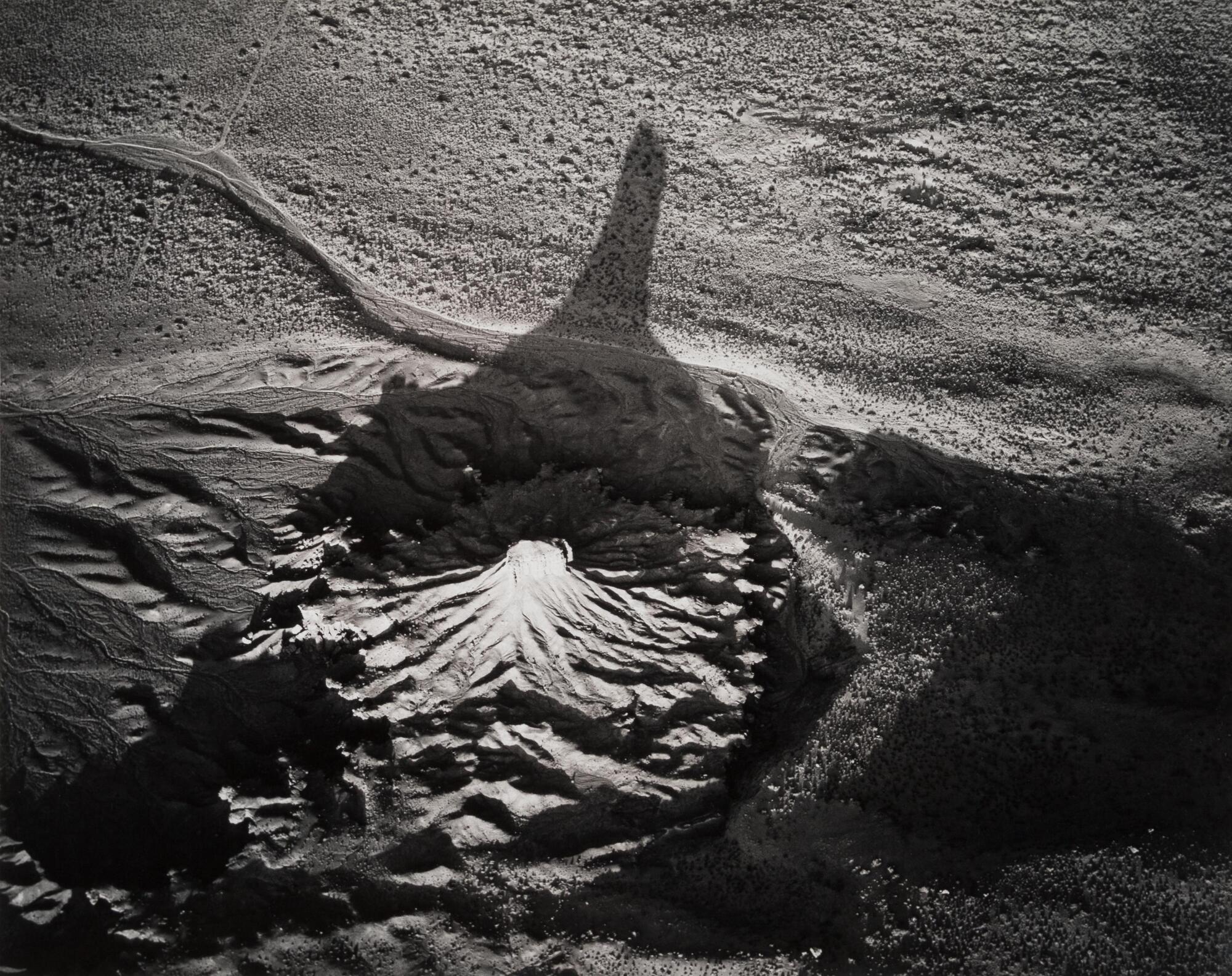 This photograph depicts an aerial view of a spire rock structure casting a long shadow on the surrounding desert.