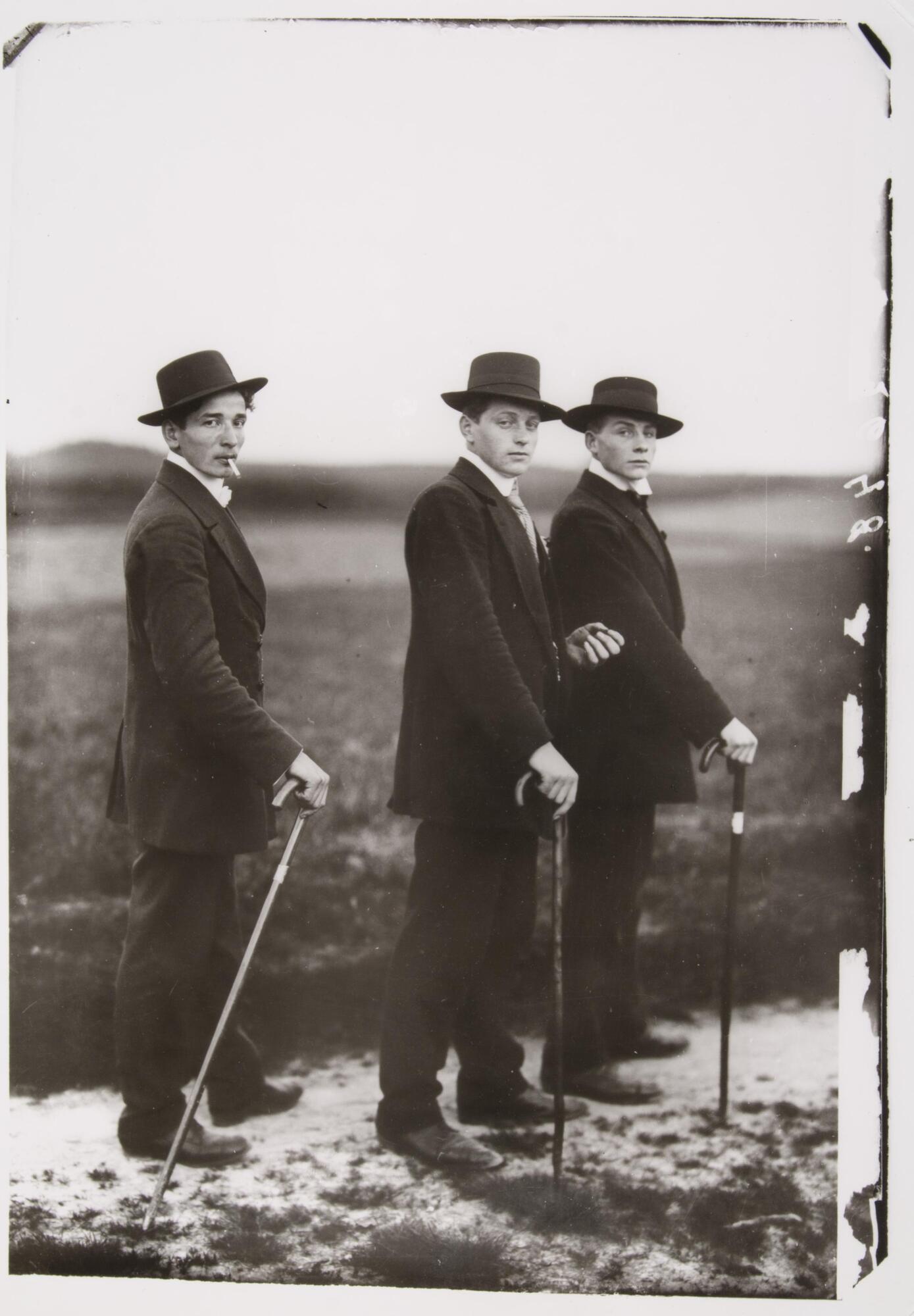 This photographic portrait of three young men shows them dressed in suits, paused during a walk on a rural road.