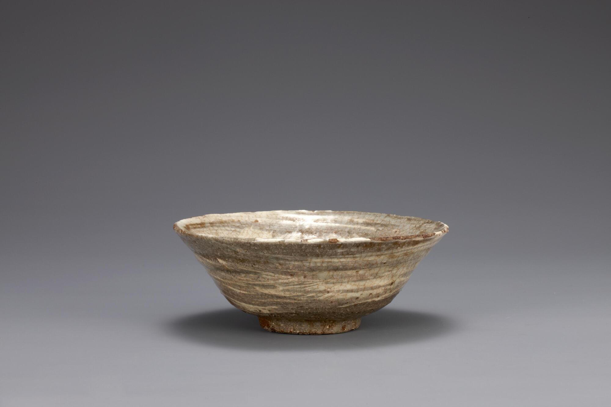 It has a flared mouth. The side is tapering gently down to the base. It was brushed with white slip interior and outerior sides.<br />
<br />
Bowls of this kind were commonly used as burial accessories in the 15th century Joseon; many have been excavated. This bowl has been rapidly spun and brush-painted with white slip on its inner and outer surfaces. The large amount of sand mixed with the clay has produced a rough texture. Traces of clay supports remain on both the inner base and the rim of foot, indicating that this bowl was stacked among other bowls while separated by clay spurs during firing. The glaze was fused well, producing a glossy surface.<br />
[Korean Collection, University of Michigan Museum of Art (2014) p.155]