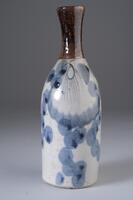 Small elongated vase with a slim body and narrow neck. Neck is a shiny brown glaze. The lower half is white with blue circular designs.