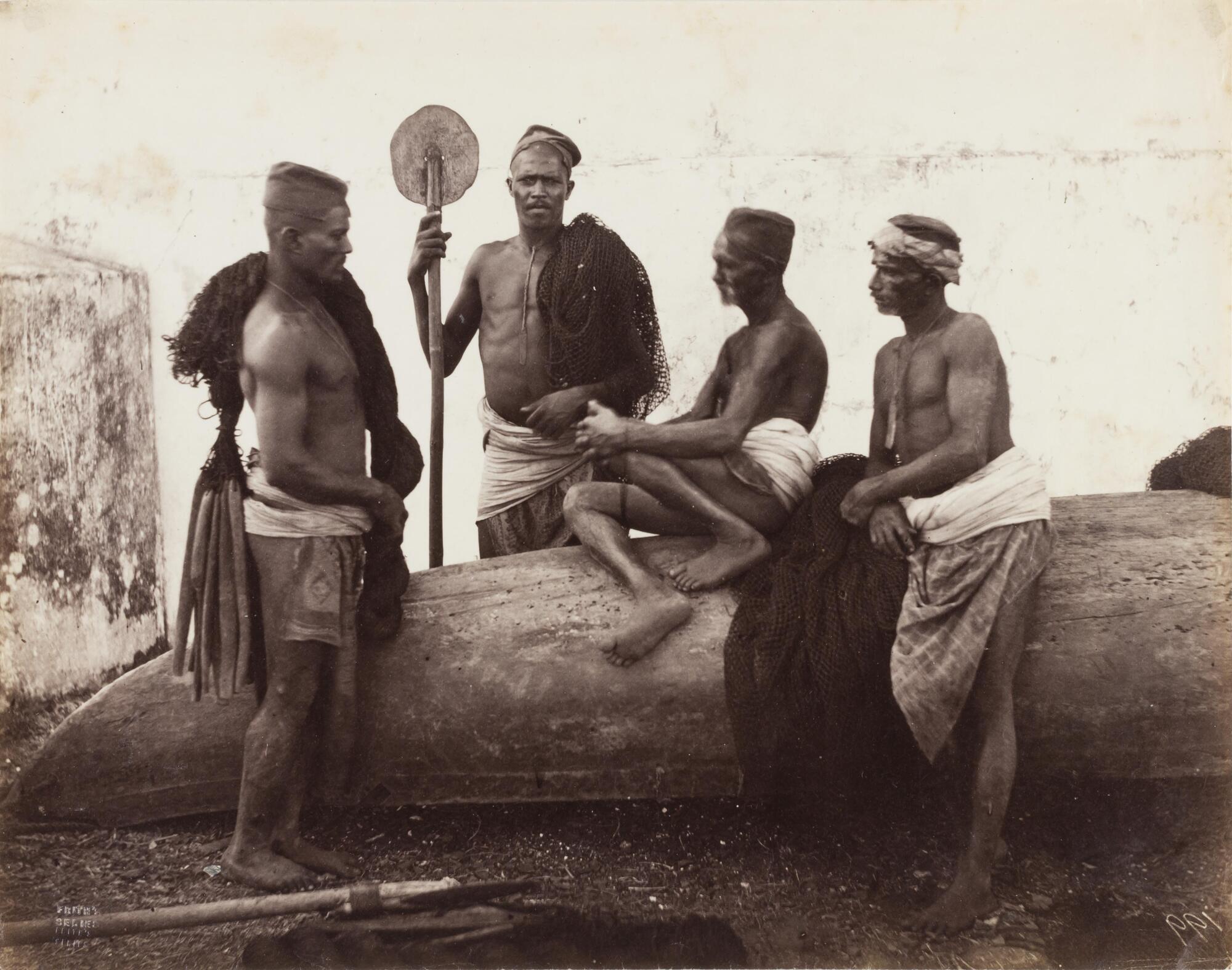 A photograph of four men sitting on and standing around an over-turned canoe. The figures hold fishing nets. One figure holds a fan and faces the viewer.