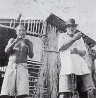 This photograph depicts two fishermen mending a large hanging net. The net stands between them and the photographer. Behind the fishermen is a wooden shack.