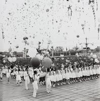 Rows of young girls in white dresses march in file in the lower foreground of the photograph while dozens of balloons float in the sky above them.