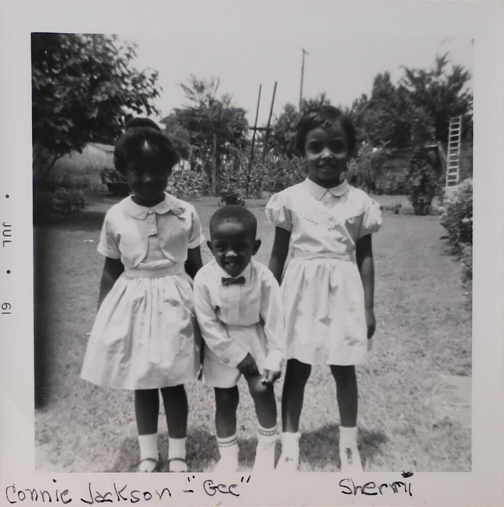 Two young girls and one young boy standing in a grassy yard. The girls are wearing light-colored knee-length dresses with collars. The boy is standing between them wearing shorts and a bow tie. He is leaning forward to grab the hem of his shorts.
