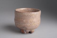 A small, ceramic saki cup with three feet at the bottom. The cup is a reddish brown color.