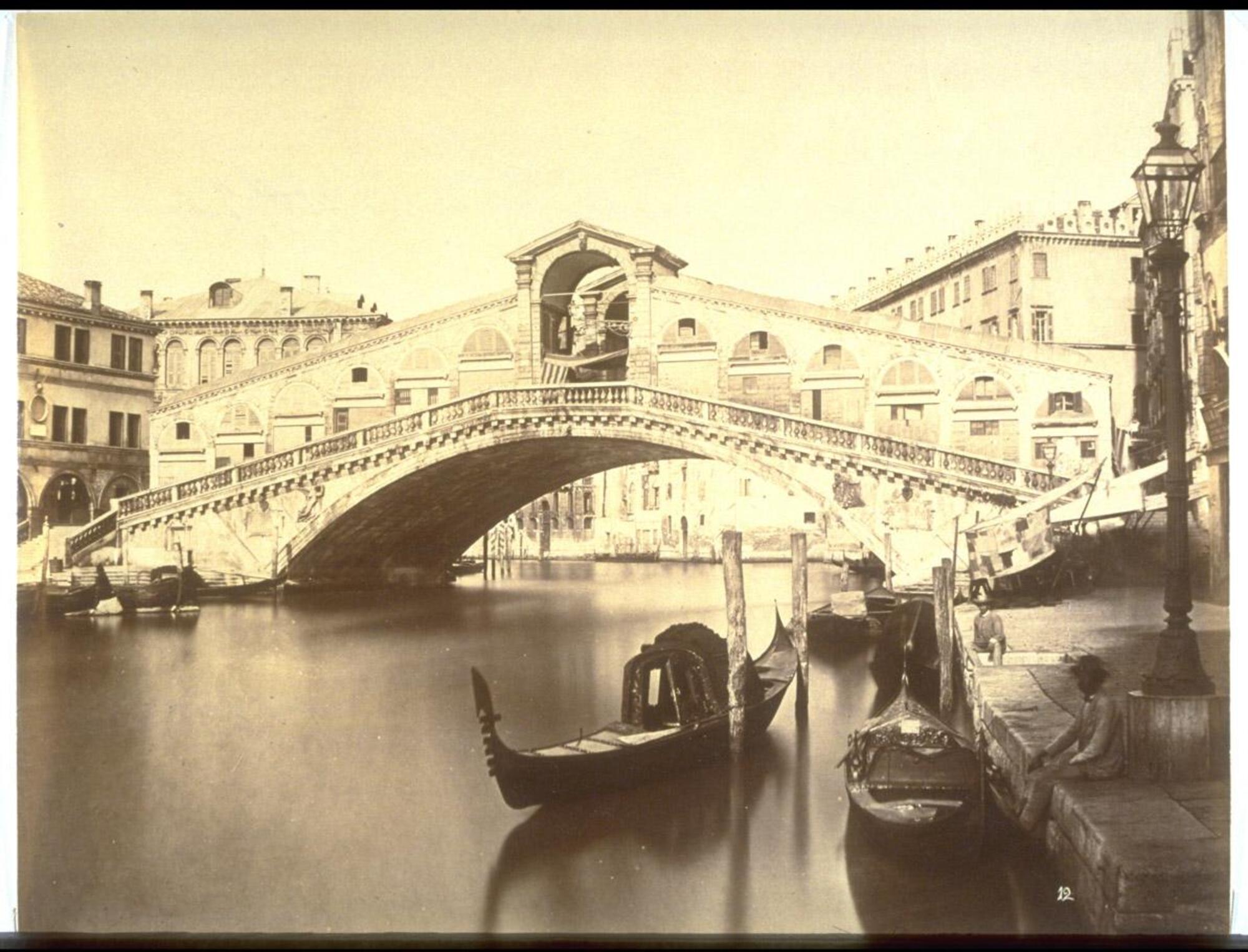 This photograph depicts a view of a marble covered bridge spanning the width of a city canal.  Along the embankments are a series of gondolas and a few seated men.