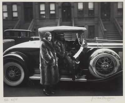 This is a photograph of an African American couple. A man sits inside his car, while a woman stands just outside, as if she is about to sit in the passenger seat. They both wear large raccoon coats. 