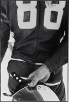 A photograph of a man's back. He holds a motorcycle helmet in his right hand behind his back and wears a leather jacket with the numbers 88.