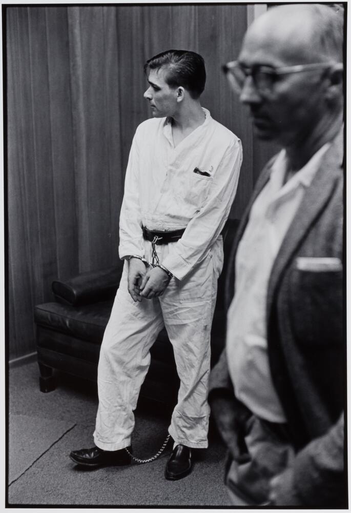 A prisoner in uniform with his hands cuffed and in front of him against a wood paneled wall. There is a man in a suit and glasses standing next to him.