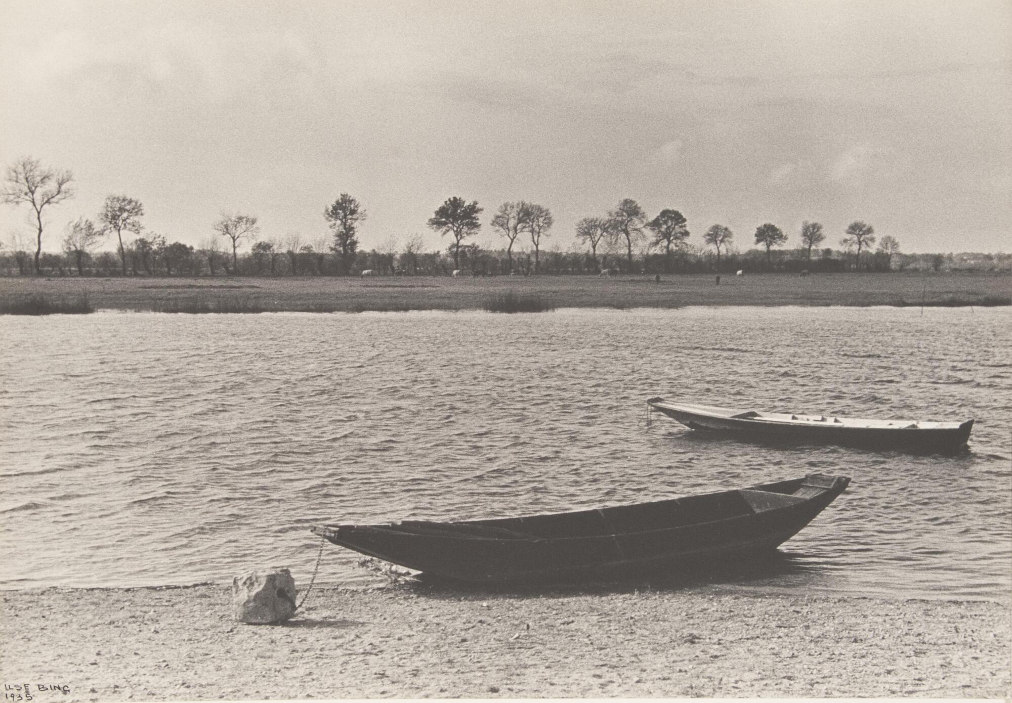 Two small boats tethered in the water and on shore of a river. The opposite tree-lined bank is also seen.