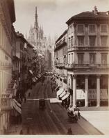 Photographed from the upper stories of a building and looking down, this image depicts a street scene in the Italian city of Milan; the city’s cathedral visible in the distance.