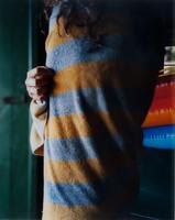 This photograph depicts a person’s torso wrapped in an blue and orange towel. The person is standing inside a dark, wooden cabin. This image is the central panel of a triptych. 