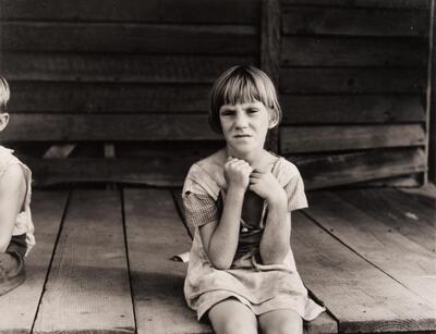 A young girl seated on a wooden porch, hands up to her neck.
