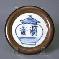 Porcelain plate with raised brown rim.  The inner circle uses blue underglaze painting to illustrate a jar with a lid that reads in kanji "fragrant orchid"
