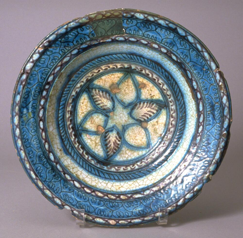 This dish contains incised floral decoration and was made with white slip with purple and turquoise underglaze, and finally glazed in a very light transparent blue. The bowl has been glued together in three pieces around the rim, with restoration also evident around the rim. 