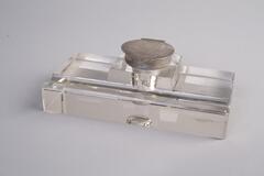 An inkwell made of glass and crystal has a rectangular shape transparent body and a metel lid cover.