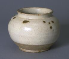 A small stoneware globular jar on a wide footring and short straight neck, covered in a white glaze that stops high above the foot ring, with dark iron-oxide dots on the shoulder. 