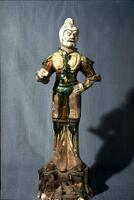 This is an earthenware standing figure of a military official or warrior. He wears Tang dynasty styled armor including a helmet, elbow-length gauntlets, a cuirass with plaques, and taces, worn over a long tunic, loose pants, and boots.  His arm is raised to hold a weapon, and he stands on a rock-styled base, which is covered in amber, green, and cream glazes. The head of the figure is unglazed with traces of mineral pigment. 