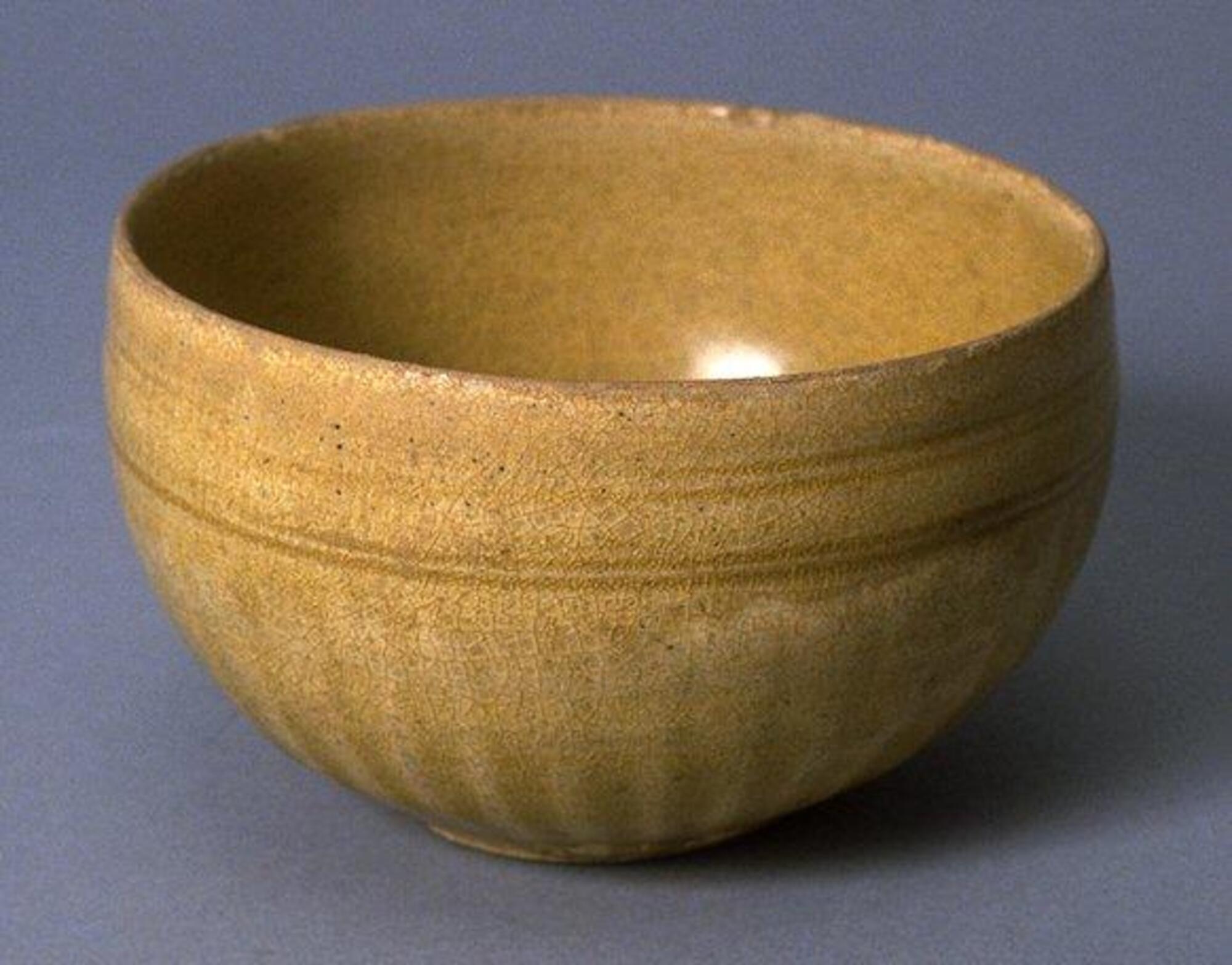 A rounded bowl, its lower two-thirds carved as a lotus blossom centered on the foot of the bowl, with two bands of double incised lines around the rim area. The pale yellow-brown glaze is worn in some areas. Five spur marks inside.