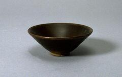 A conical bowl on a tall straight foot ring, covered in a dark brown-black mottled glaze, thinning to brown at the rim.