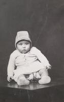 A portrait of a baby in a bonnet and matching outfit seated on a table. The infant is supported by a wire holder, and leans to the left of the frame. 