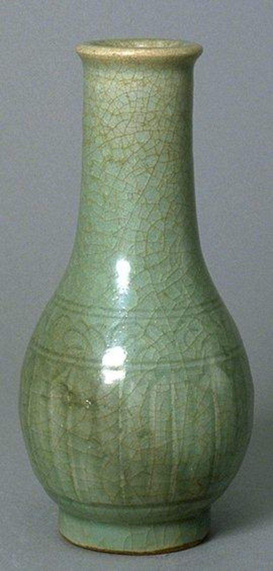 A stoneware bottle-shaped vase with a bulbous body tapering to a tall narrow neck with everted rim, on a tall footring. It has incised lappets around the lower body and a band of floral meander around the shoulder. It is covered in a craqueleur celadon glaze. 