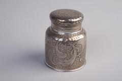 Silver inkwell made with glass, metal and silver; it has a cylinder body and a round top lid. The body of the inkwell is covered with patterns.