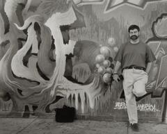 A man holding a camera, wearing khakis, one leg up against a graffiti covered wall.