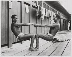 Photograph depicting two men engaged in calisthenics, using their joined hands and connected knees to pull away from each other. In the background a man lies on the same wooden deck, while another man clothed in a towel can be seen in the background of the image.
