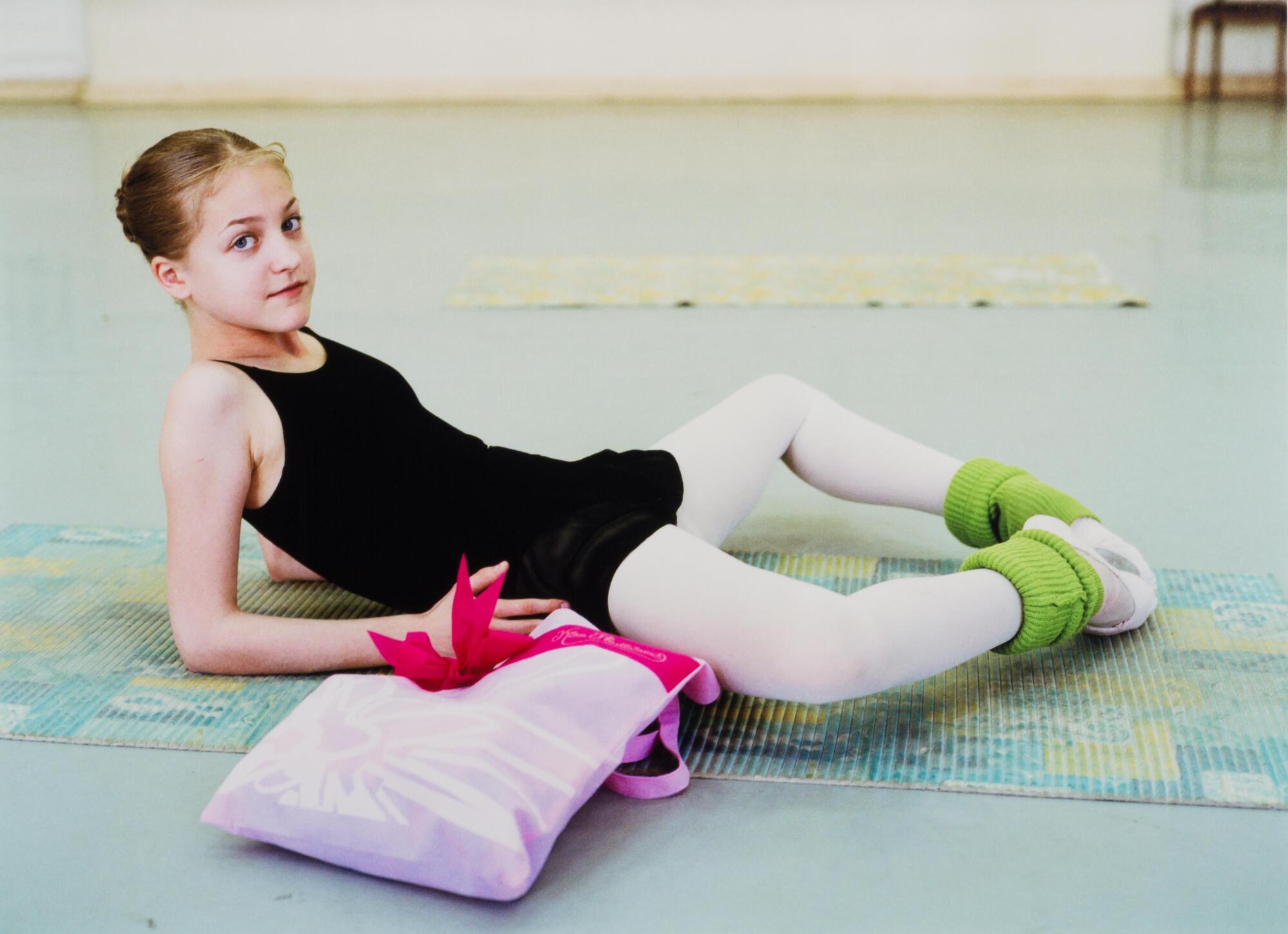 A girl in a black leotard, pink tights and green leg warmers lounging on a light blue floor.