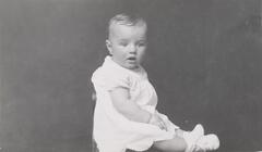 A portrait of an infant seated on a table, body facing the right side of the frame, head turned toward the camera.