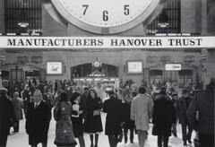 A interior view of Grand Central Terminal Concourse. Large clock and Manufacturers Hanover Trust sign in background. Very busy with many people walking around.