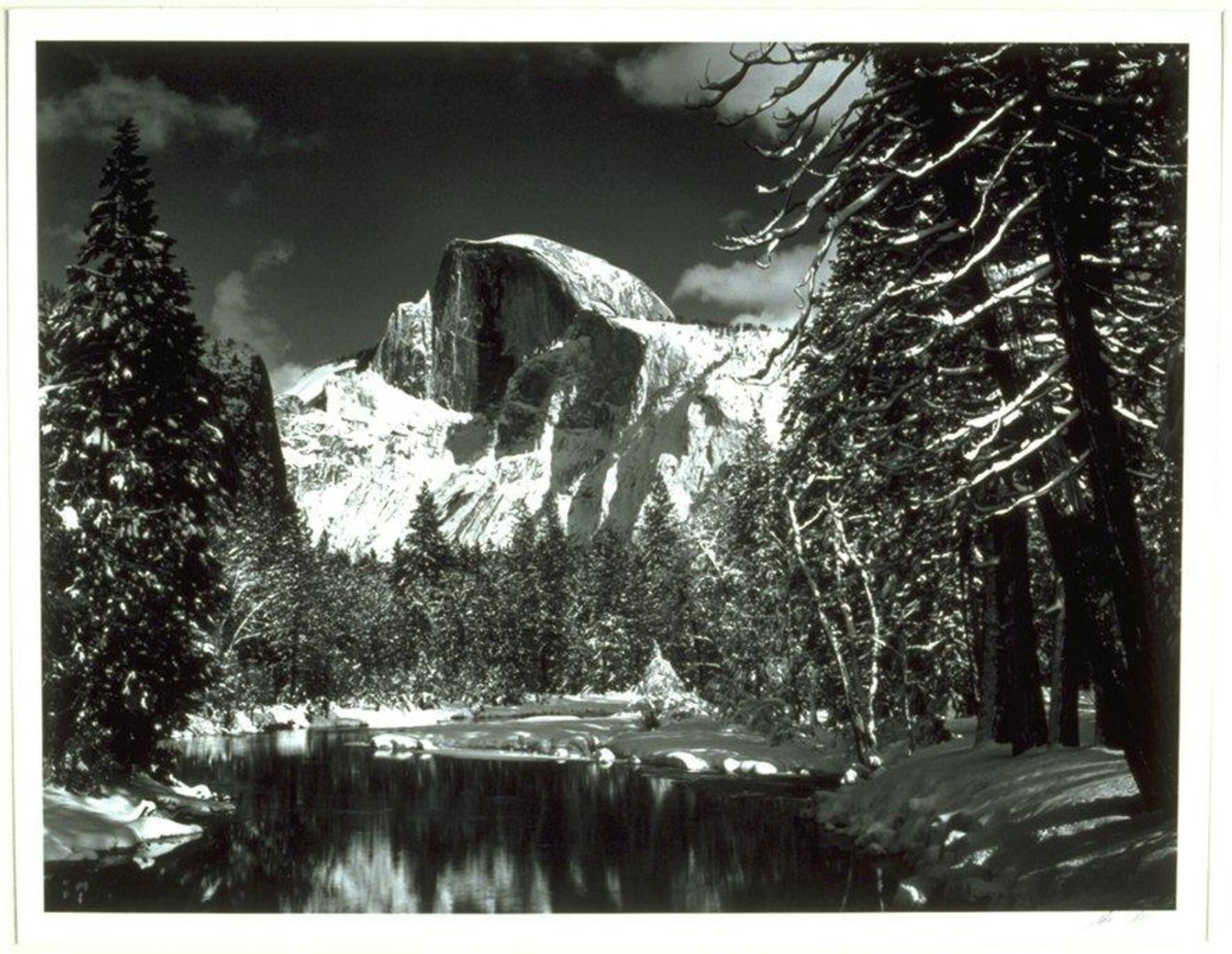 The photograph depicts a river lined with snow-covered banks and pine trees and a snow-capped mountain rising in the distance.  