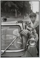 A photograph of three boys in a car. A young man sits in the driver's seat and is visible through the windshield as he reaches his hand out to adjust the windshield wipers. Another boy sits in the back seat, while on the right a boy leans against the exterior of the car.