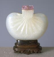 A white nephrite jade snuff bottle that is shaped like a handbag. It has a rounded bottom and shoulders. Where the clasp at the top of the handbag would be is a square opening of the snuff bottle that is capped with transparent pink cap. On this opening are carved vertical lines that continue onto the surface of the snuff bottle to replicate creases. Also decorated on the surface are carved abstract curls. The snuff bottle is sitting on a wooden stand.